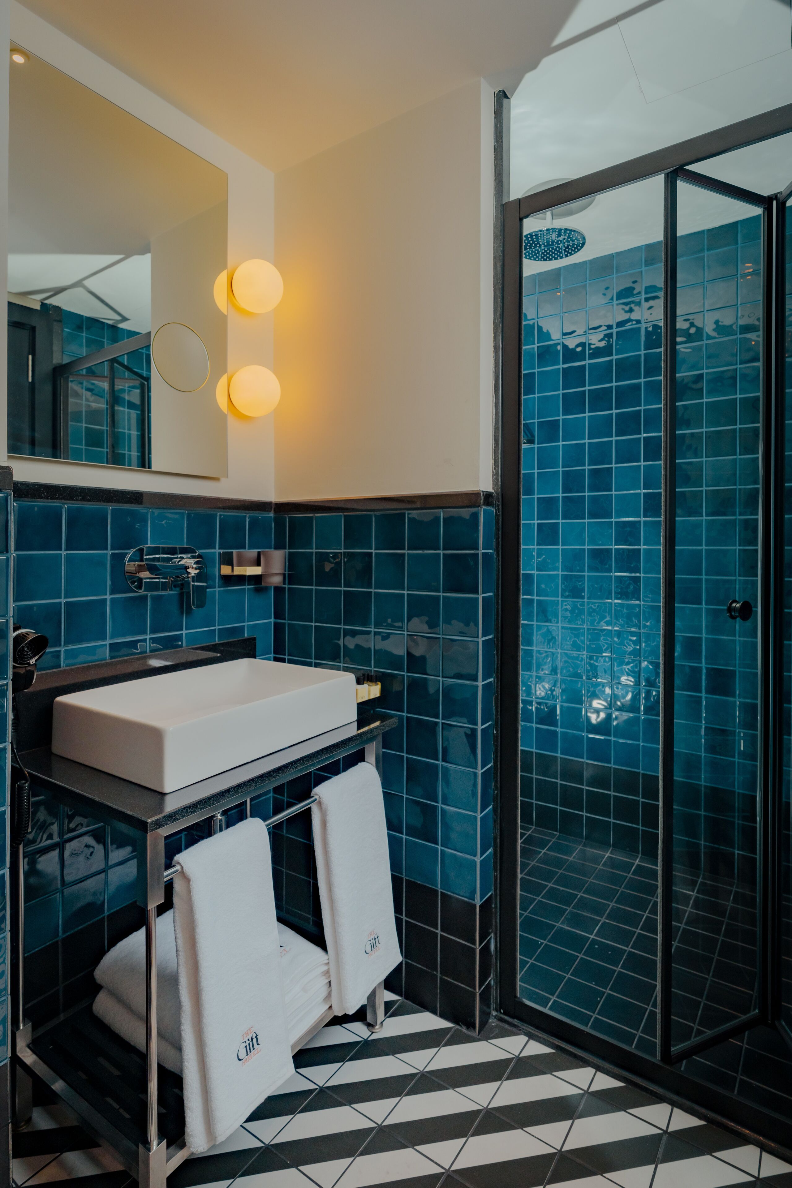 Un baño moderno con un lavamanos blanco, azulejos azules, cabina de ducha de vidrio y piso a cuadros blanco y negro.