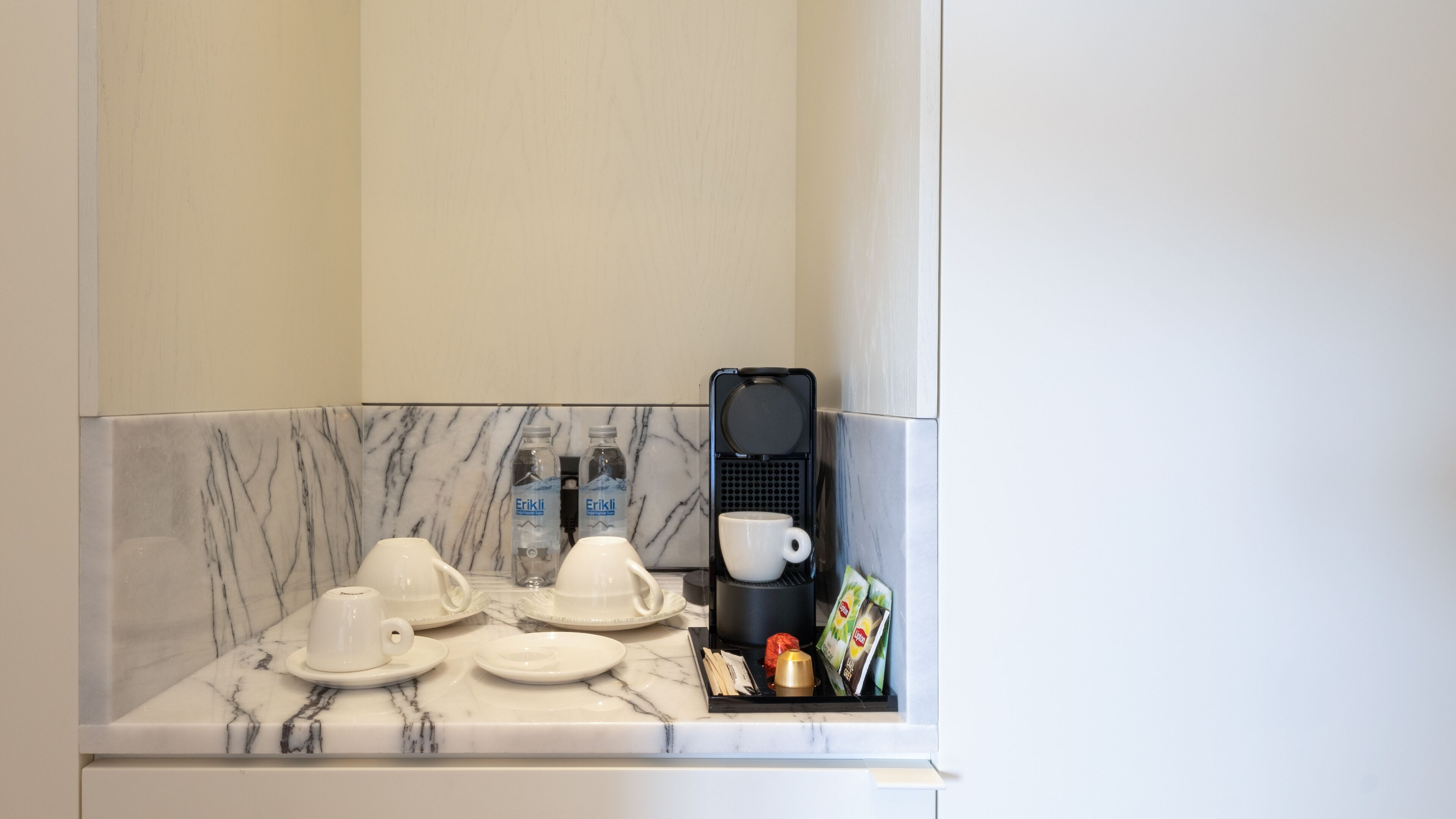 A compact kitchenette with a coffee maker, two cups, a bottle of water, and a small tray with condiments.