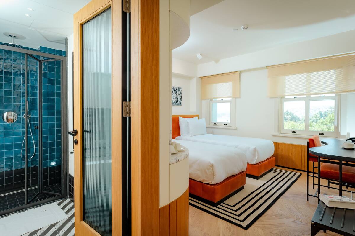 A modern hotel room with a glass shower enclosure, a bed with orange headboard, a dining area with a table and chairs, and a large window with blinds.