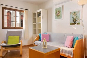 Un salon comprenant un fauteuil à bascule en bois, un canapé gris agrémenté de coussins colorés, un meuble blanc et une petite table sur laquelle est posée une lanterne.