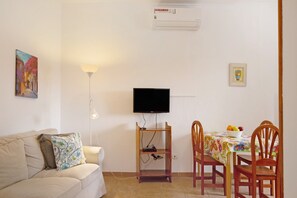 A living room with a sofa, a TV mounted on a stand, a dining table with chairs, and a wall-mounted air conditioner.