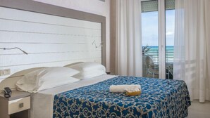 Una habitación de hotel con cama, mesita de noche, ventana con cortinas y vista al mar.