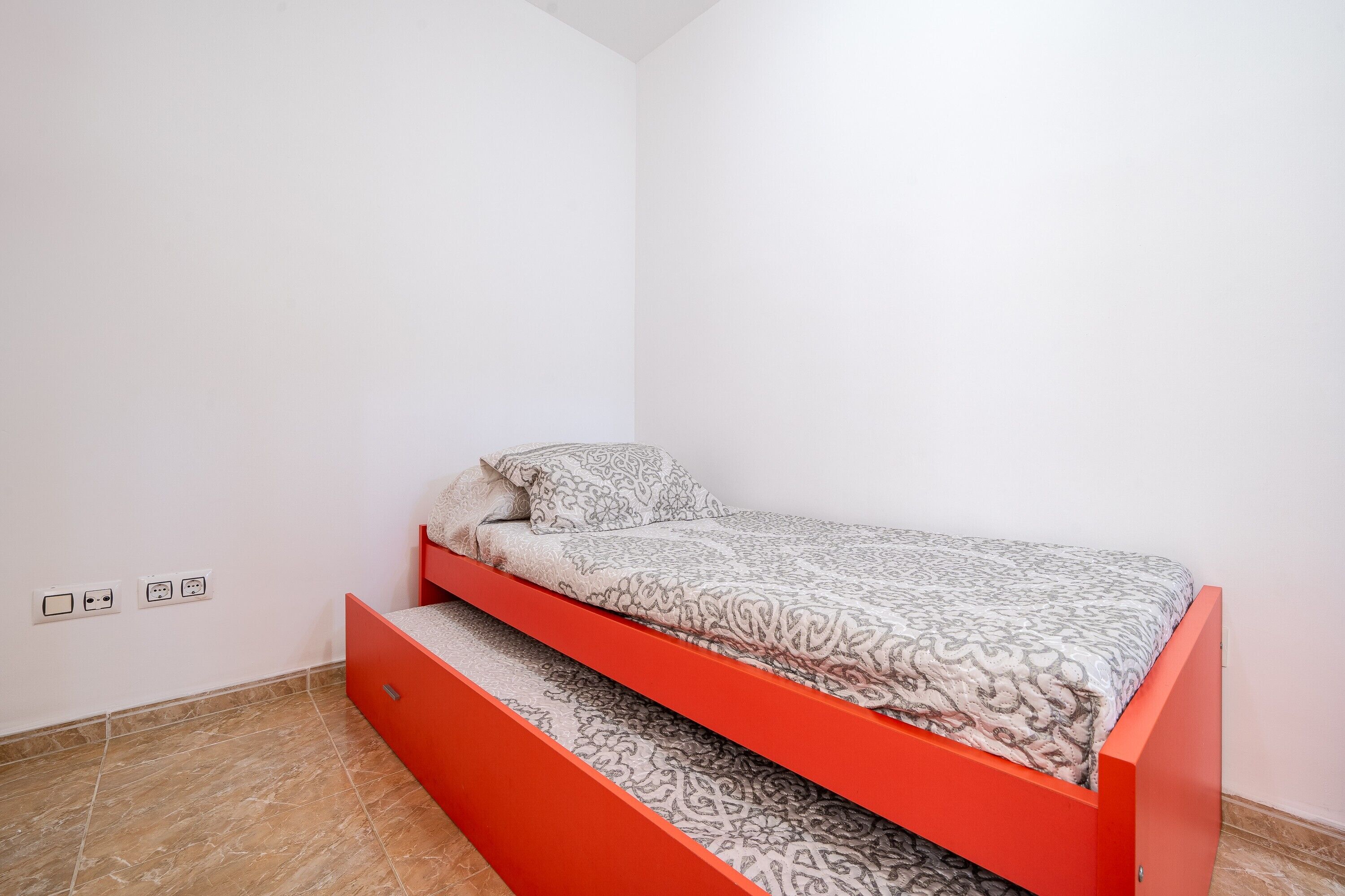 Une petite chambre avec un lit simple, un cadre de lit rouge, une literie à motifs et un sol carrelé.