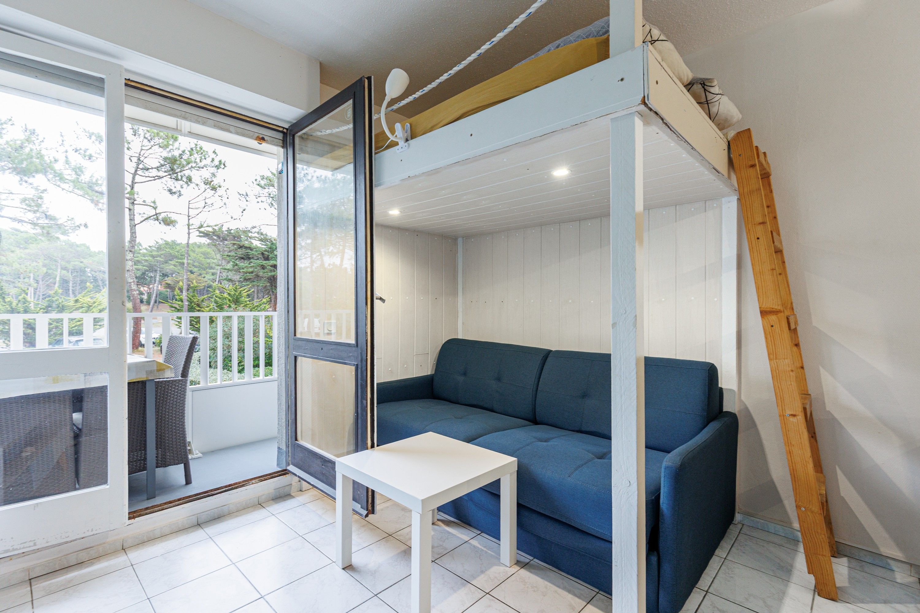 Una habitación con un sofá azul, una mesita blanca y un balcón con una silla.