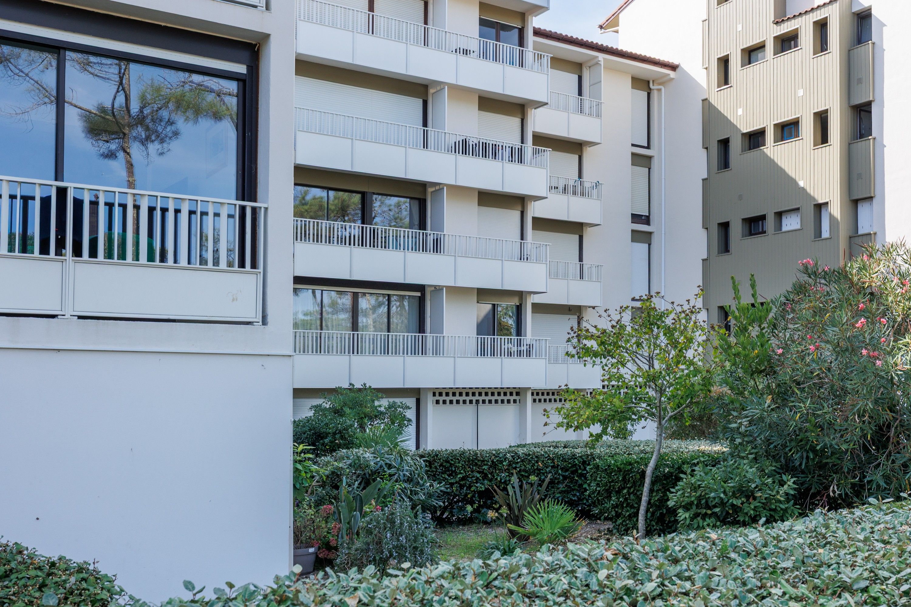 Un edificio residencial de varios pisos con balcones, un jardín con diversas plantas y un cielo despejado.