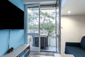 Een kamer met een televisie aan de muur, een bank, een tafel en een schuifdeur die uitkomt op een terras met een balkon en uitzicht op bomen en geparkeerde auto's.