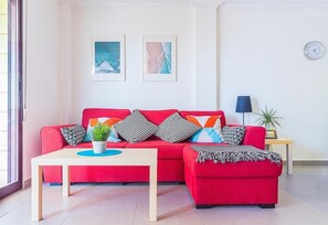 Un canapé rouge, une petite table et une plante, une horloge au mur et une lampe sur une table d’appoint.