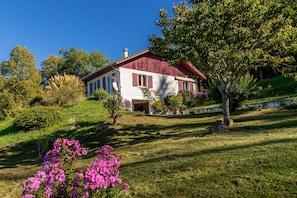 Une maison avec un toit rouge, des murs blancs et un jardin où poussent des fleurs roses.