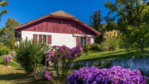 Een rode schuurwoning met een schuin dak, omgeven door weelderige begroeiing en paarse bloemen.