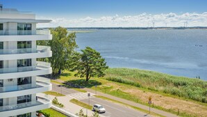 Un edificio moderno con varios balcones que dan a una masa de agua, una carretera con un cartel de límite de velocidad y aerogeneradores a lo lejos.