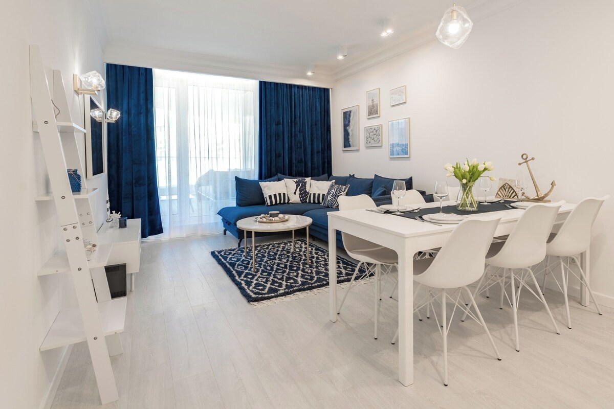 Un salon moderne avec un canapé, une table à manger et des chaises. On y trouve une grande fenêtre avec des rideaux, un mur blanc orné de tableaux et un plancher de couleur claire.