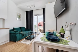 Un salon moderne avec un canapé vert, une table à manger dressée avec de l’assiette et des couverts, un téléviseur à écran plat fixé au mur et une vue sur l’extérieur grâce à une grande fenêtre.