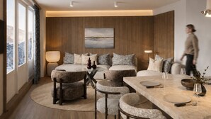 A modern hotel room with a wooden panel wall, a large bed with patterned pillows, a glass coffee table, and a dining area with a marble table.