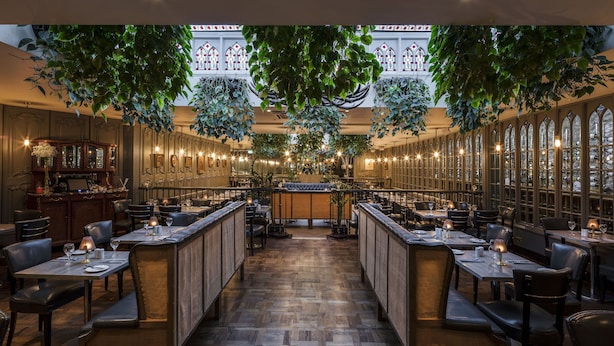 A restaurant with wooden flooring, high ceilings, and hanging plants. Tables are set with candles and wine glasses.
