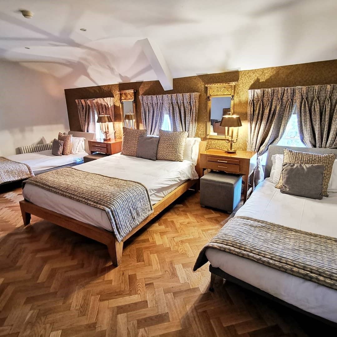 Quarto de hotel com uma cama grande, duas camas menores, piso de madeira em padrão espinha de peixe e um espelho sobre as camas menores.