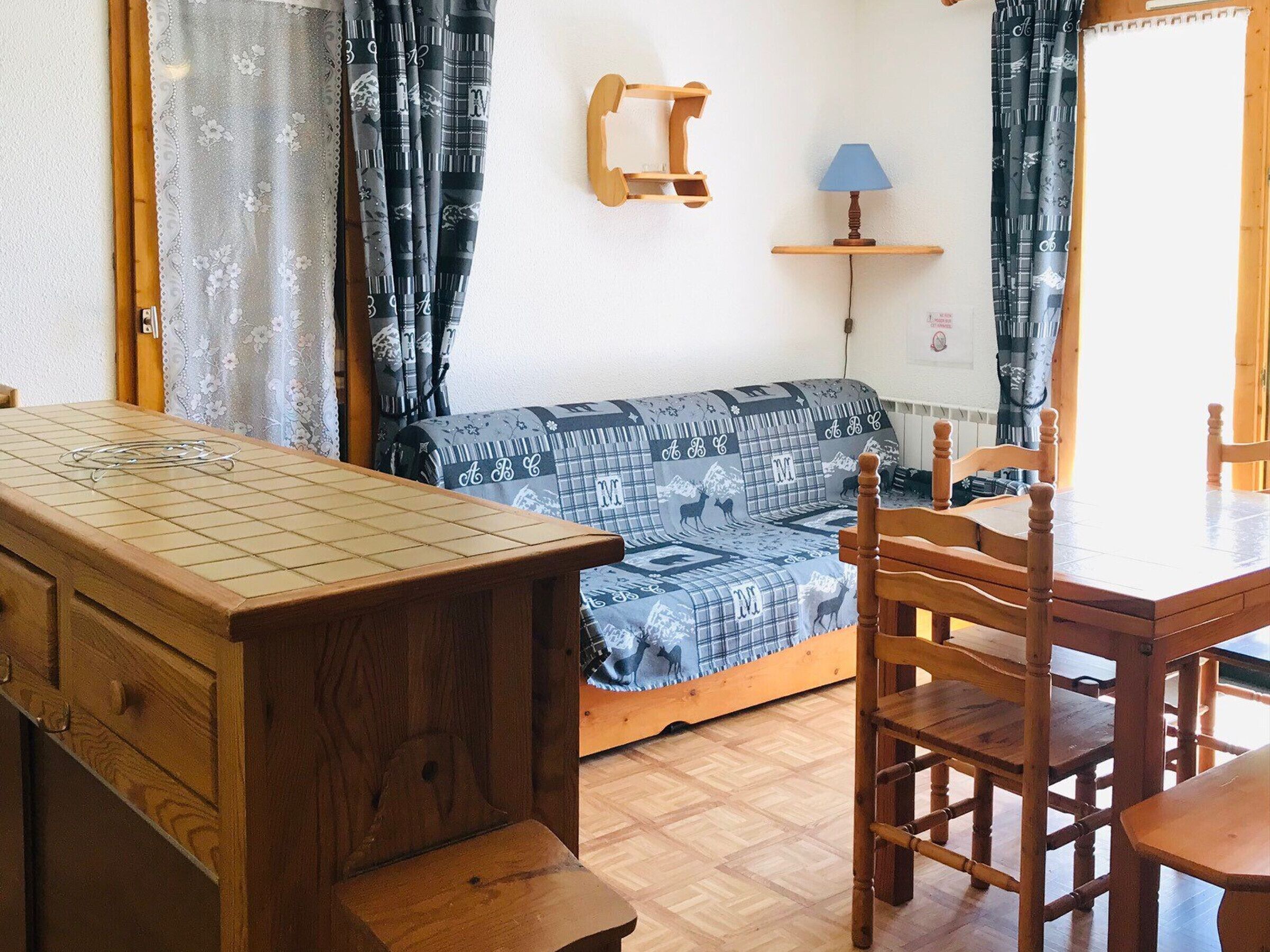 Una habitación acogedora con una mesa de madera, un sofá con cojines estampados, una lámpara en un estante pequeño y una ventana con cortinas.