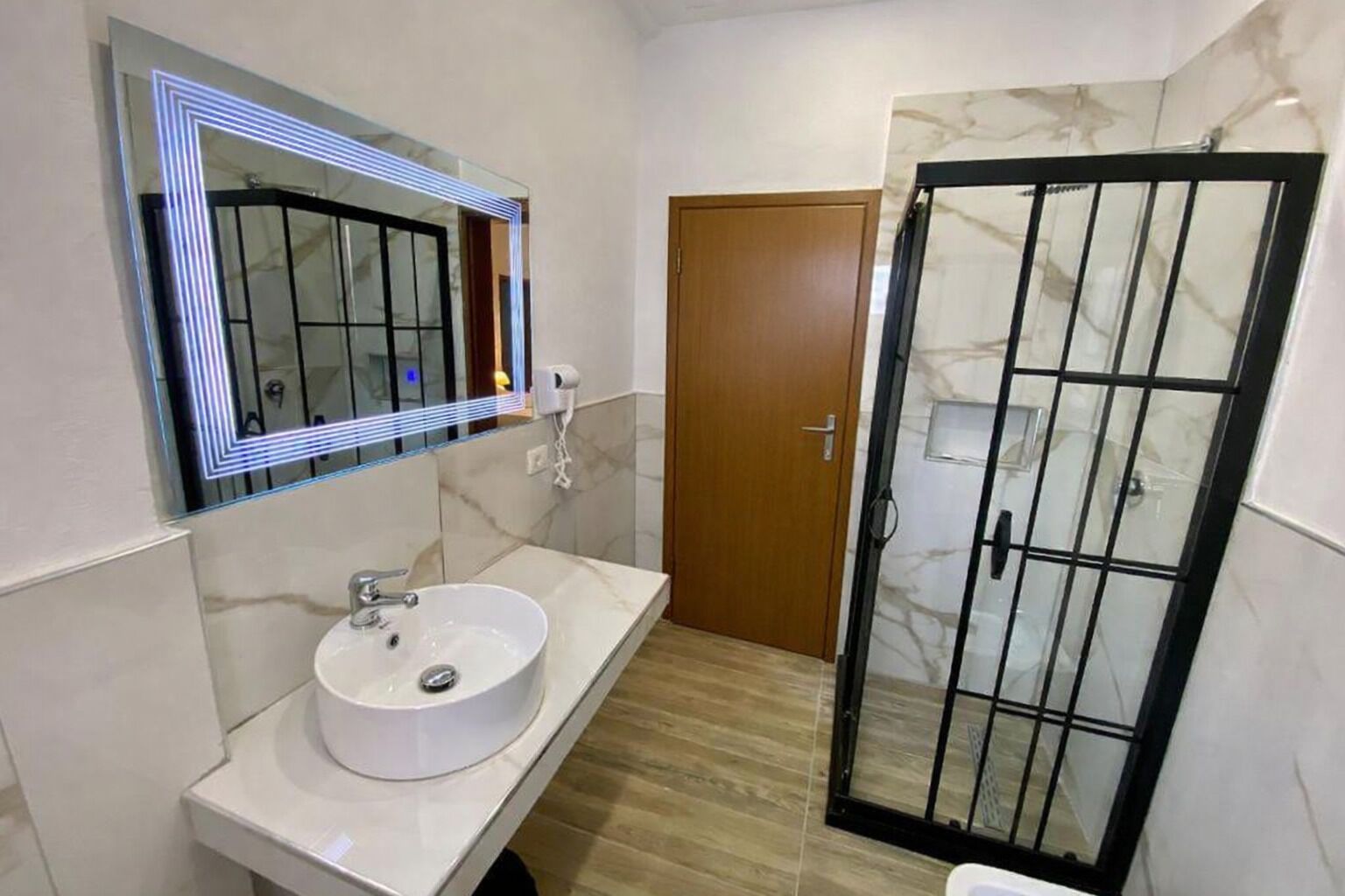 Un baño con un lavamanos blanco, un espejo con luces LED, una puerta de madera y un box de ducha de vidrio.