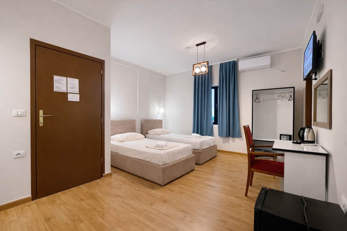 A hotel room with a wooden floor, a desk with a chair, a bed, a TV, and a window with curtains.