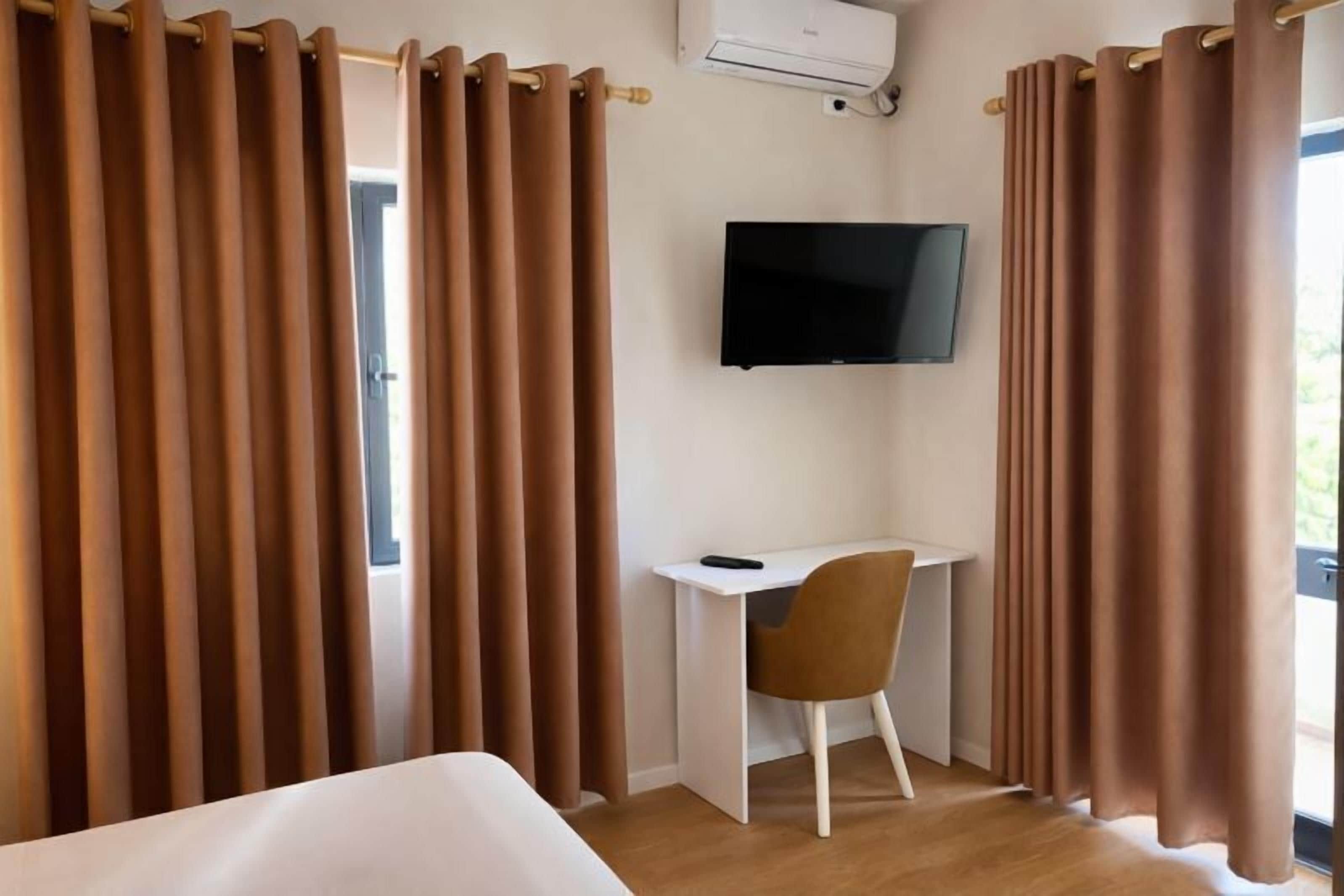 Een hotelkamer met een bed, een bureau met stoel, een wandtelevisie en bruine gordijnen.