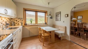 Une cuisine avec des meubles de rangement en bois, un plan de travail blanc, un évier, une fenêtre donnant sur la campagne, un coin repas avec une table et des chaises, et un radiateur.