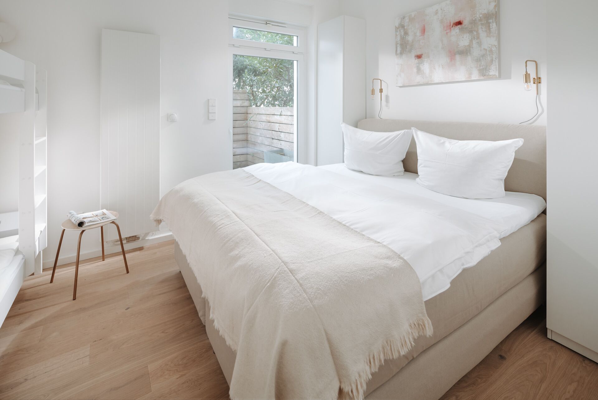 A minimalist bedroom with a large bed, a small stool, and a window with a view of trees.