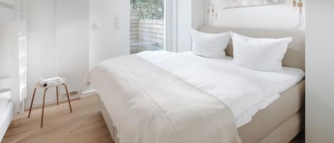 Quarto minimalista com uma cama grande, um banquinho pequeno e uma janela com vista para árvores.