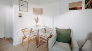 A room with a white table, two wooden chairs, a green cushion on a grey sofa, a lamp, and a framed picture on the wall. - // V I L L A F R E S E N A // Bude 3 (Norderney)