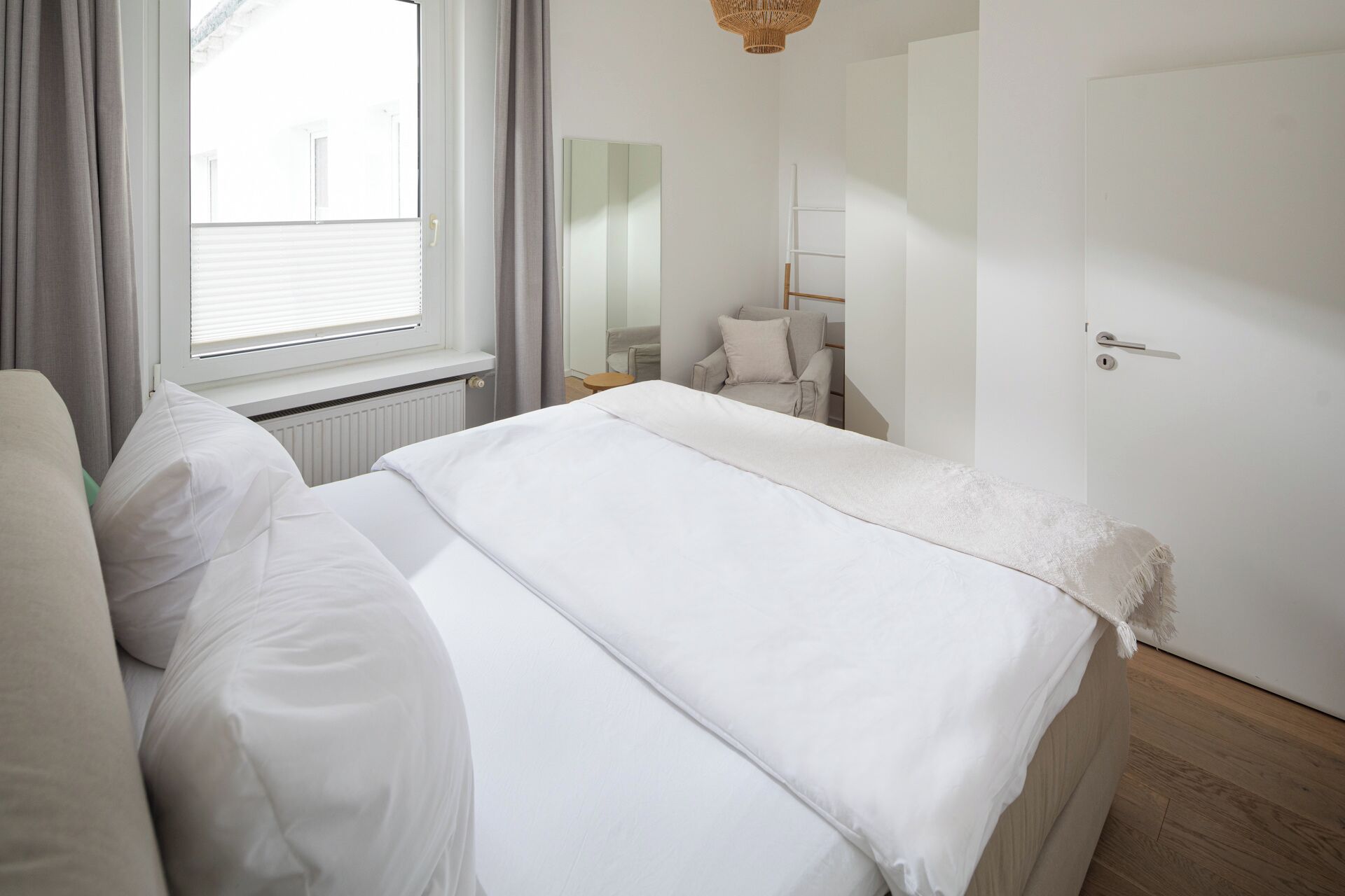 A bedroom with a bed, a window with curtains, a chair, a bedside table, and a door.