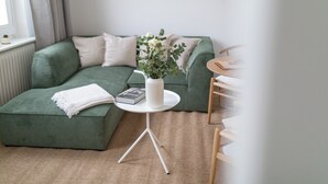 A modern living room with a green sectional sofa, a small round table with a white vase and book, and a wooden chair. - // V I L L A F R E S E N A // Bude 6 (Norderney)