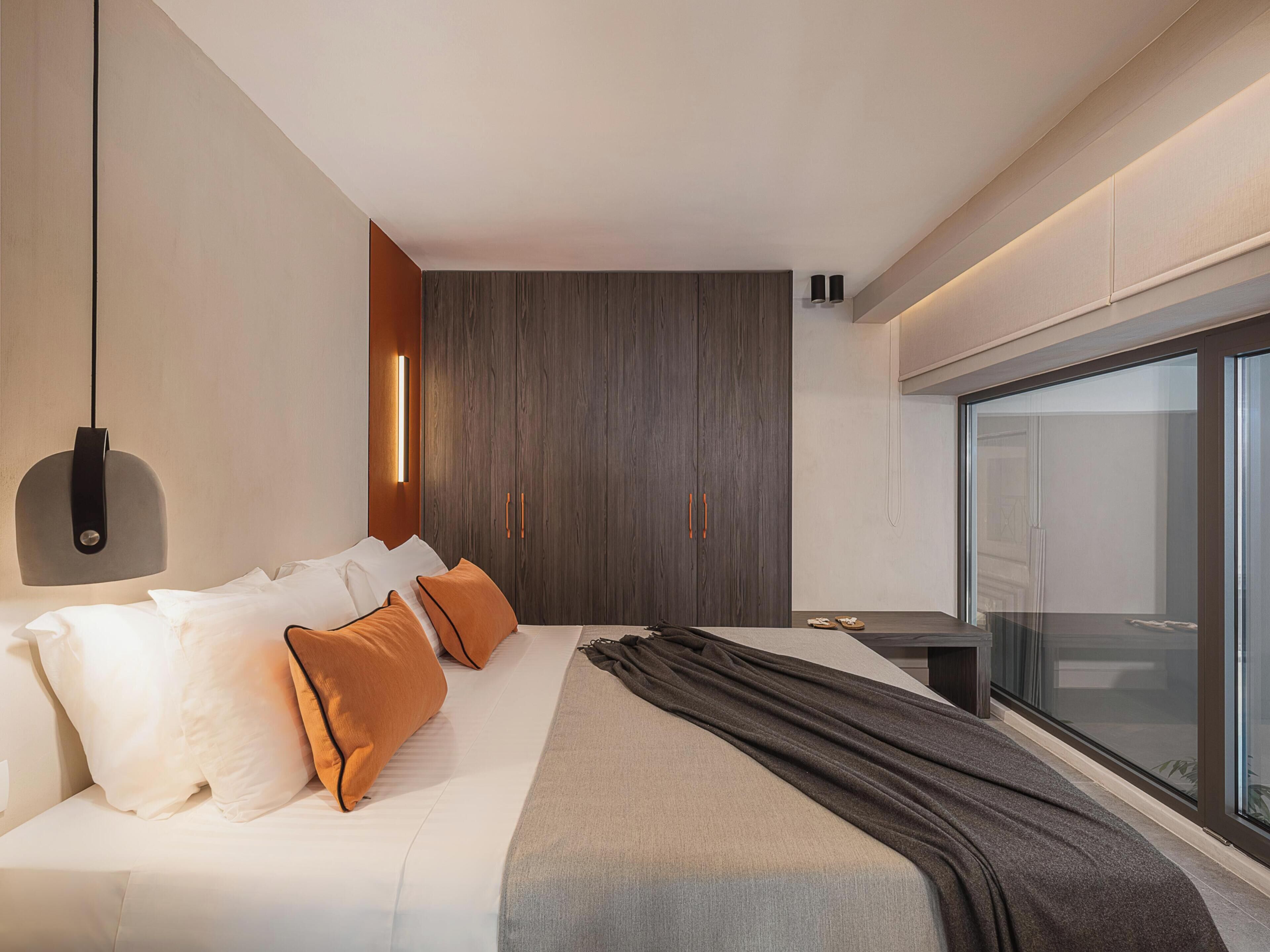 A modern bedroom with a bed, pillows, a bedside table, a wardrobe, and a large window.