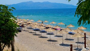 A beach with rows of thatched umbrellas and beach chairs, a clear view of the sea, and distant mountains. - Hotel Emblem (Sarandë)