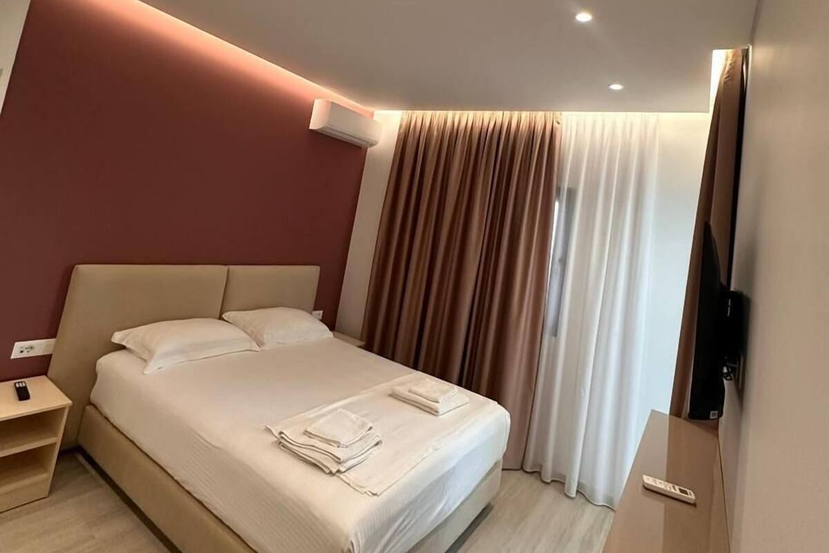 A modern hotel room with a large bed, a bedside table, an air conditioning unit, and a flat-screen TV mounted on the wall.