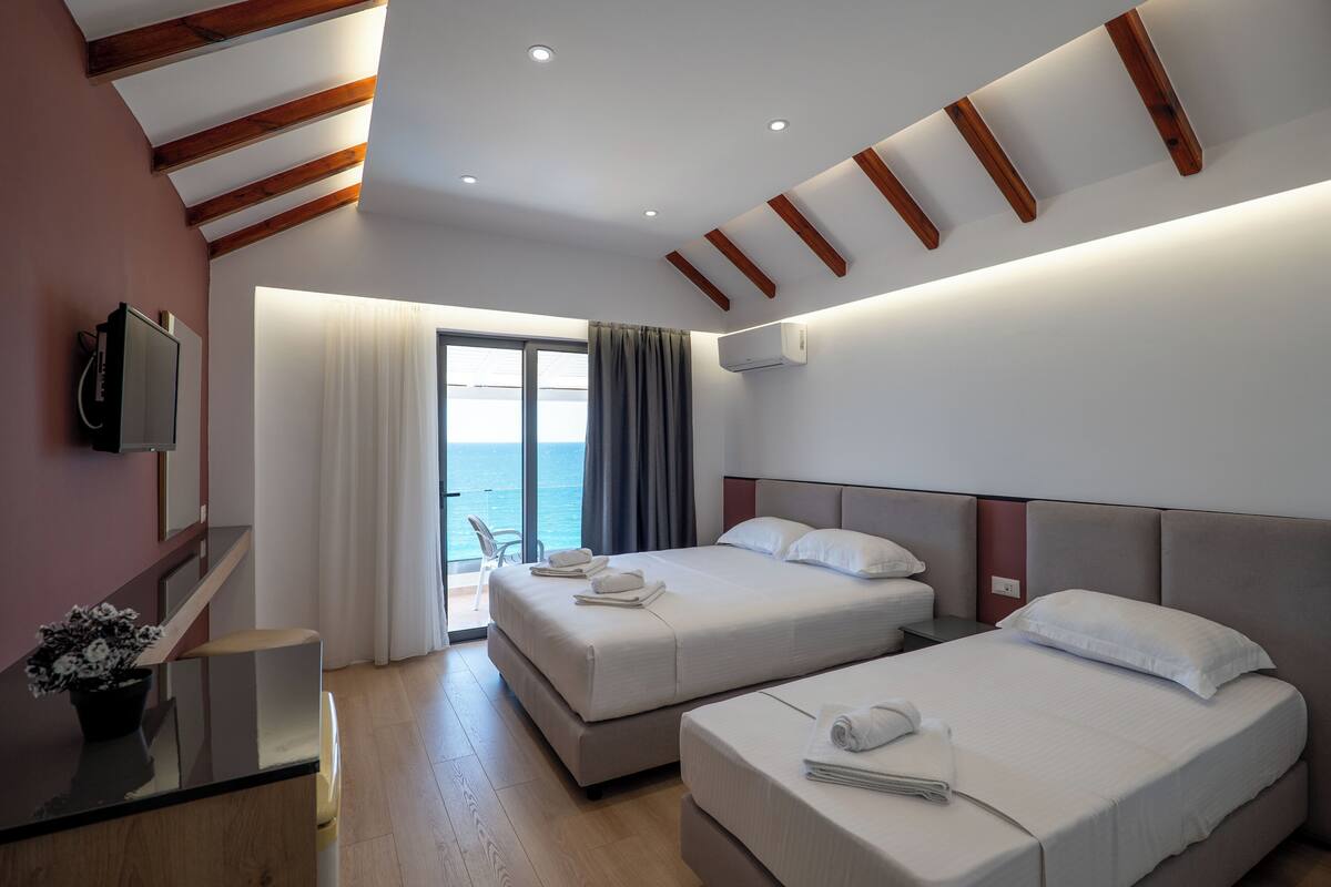 A hotel room with two beds, a TV, a desk, and a view of the sea.