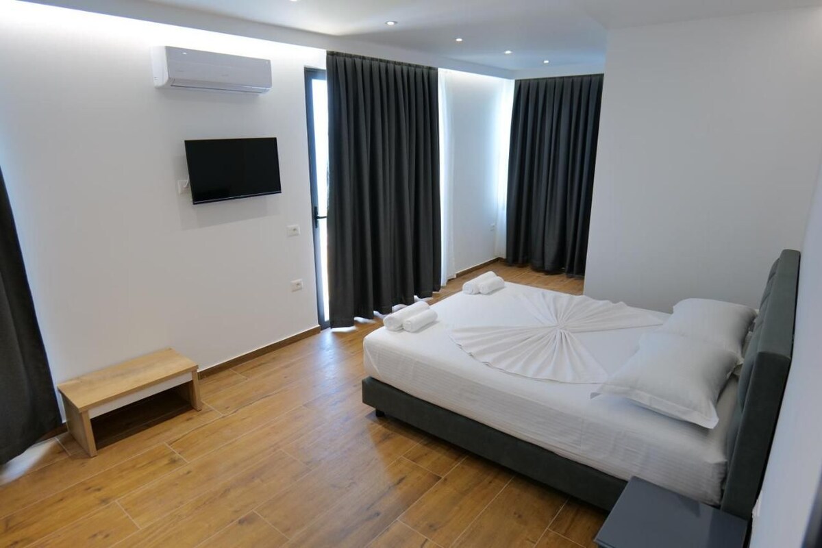 Habitación de hotel moderna con cama, televisor, pequeño banco de madera y ventana con cortinas.