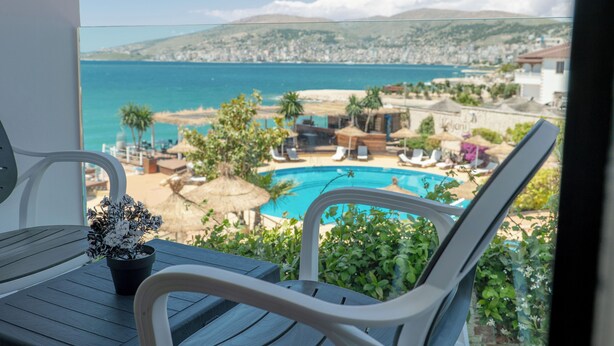 A balcony with a view of a swimming pool, lounge chairs, and a mountainous landscape. - Hotel Emblem (Sarandë)