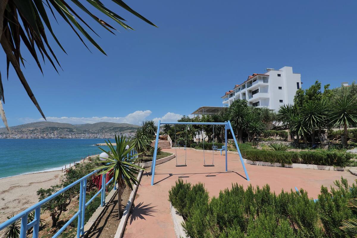 A beachside park with a swing set, a white building, and a view of the sea.