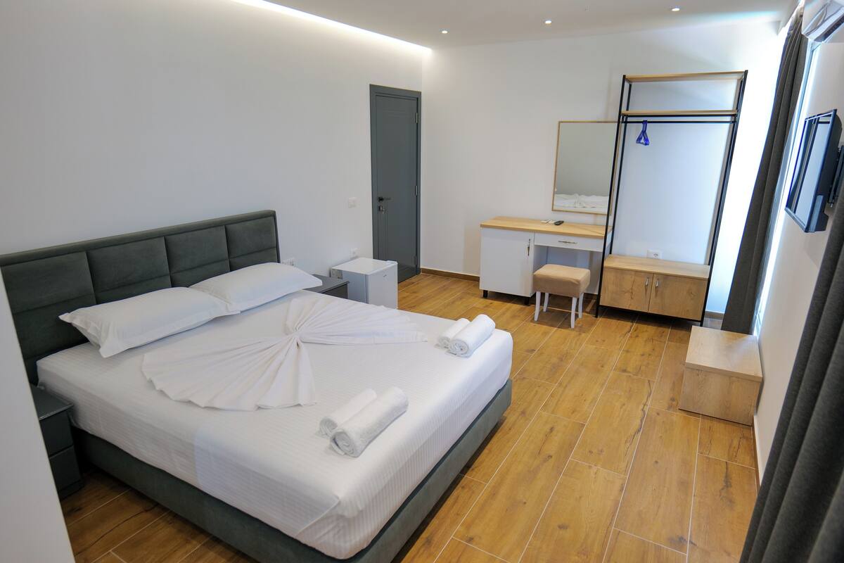 A neatly made bed with white linens and rolled towels, a wooden floor, a small bedside table, a desk with a mirror, and a wardrobe.