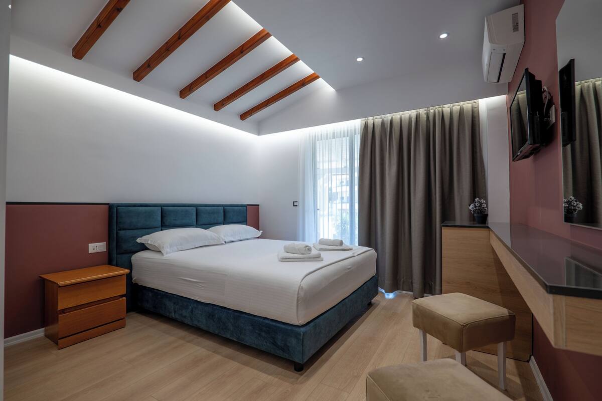 A modern bedroom with a large bed, wooden headboard, bedside tables, a bench, a TV, and a mirror.