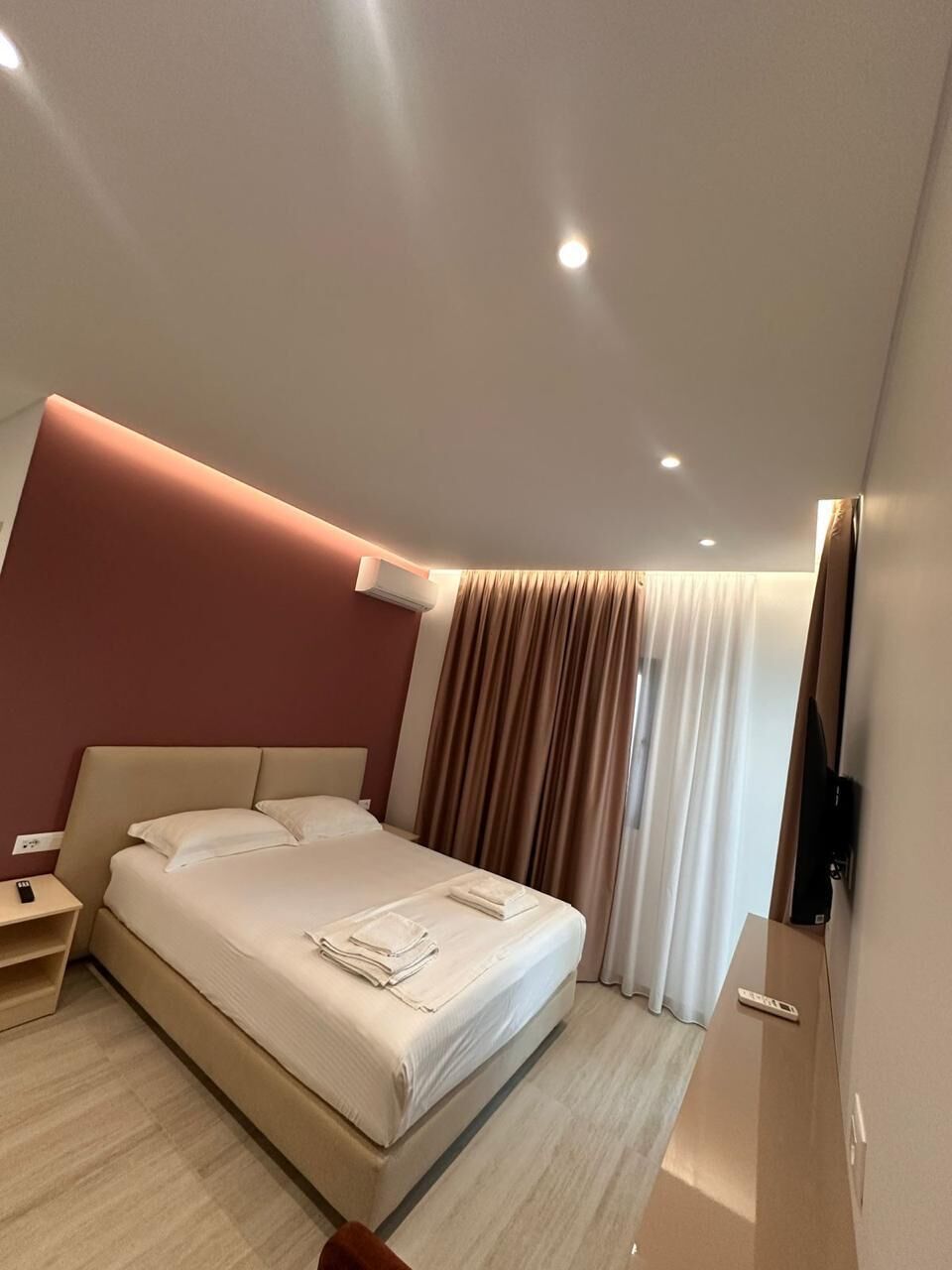 A modern hotel room with a large bed, a bedside table, an air conditioning unit, and a flat-screen TV mounted on the wall.