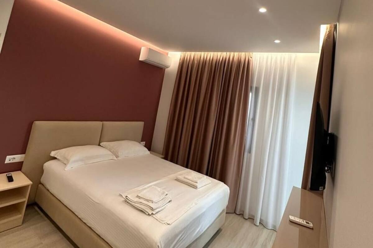 A modern hotel room with a large bed, a bedside table, an air conditioning unit, and a flat-screen TV mounted on the wall.