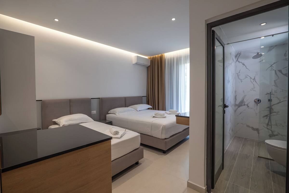 A modern hotel room with two beds, a wooden desk, and a glass-enclosed shower area.