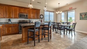A spacious kitchen with wooden cabinets, a central island with bar stools, a dining area with a table and chairs, and a large window with blinds. - Desert Escape: Sand Dunes + Golf + Resort Access (Hurricane)