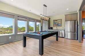 A modern kitchen with a pool table, large windows, and a stainless steel refrigerator.