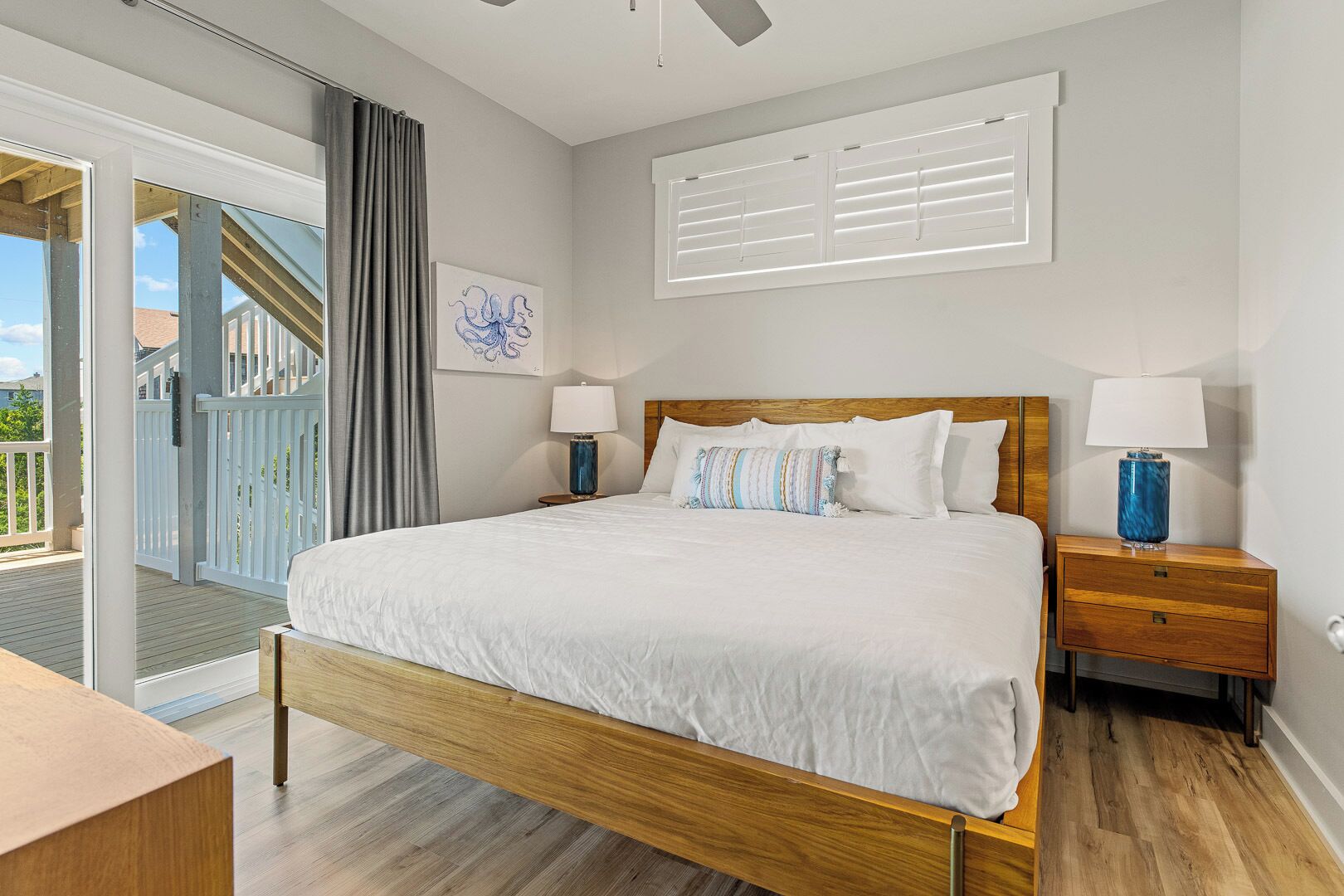 A bedroom with a wooden bed, white bedding, a nightstand with a lamp, a window with blinds, and a view of a deck and buildings outside.