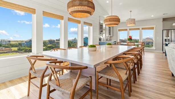 Uma ampla área de jantar com uma mesa comprida e cadeiras de vime, grandes janelas que oferecem vista do bairro circundante e uma cozinha moderna ao fundo.