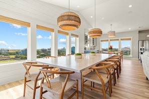 A spacious dining area with a long table and wicker chairs, large windows offering a view of the surrounding neighborhood, and a modern kitchen in the background.