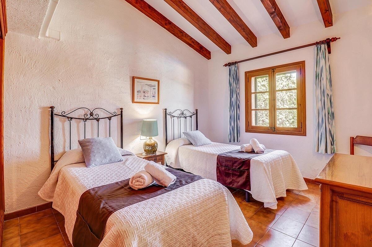 A room with two beds, wooden beams on the ceiling, a window with curtains, and a small table.