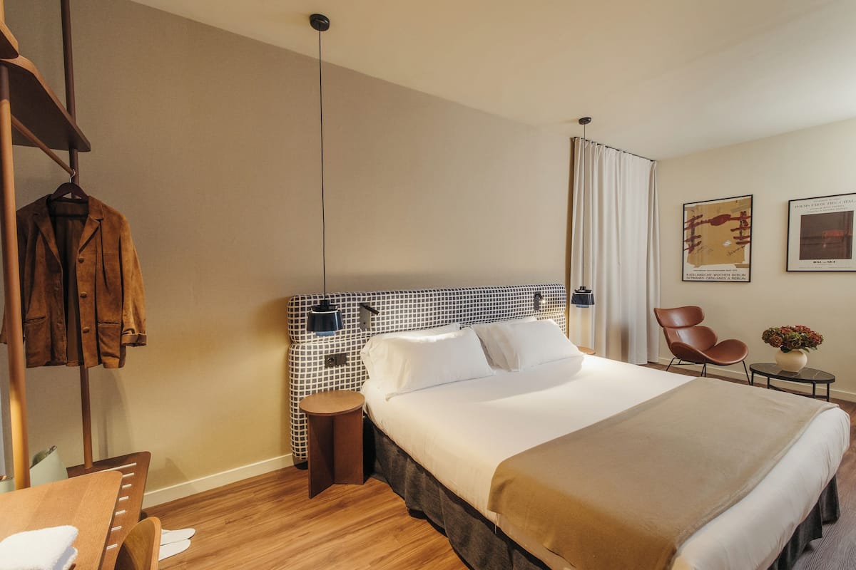 Una habitación de hotel con una cama grande, una silla de madera, una mesita pequeña y un perchero.