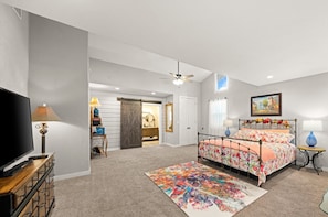 A bedroom with a large bed, a TV, a dresser, a desk, and a bathroom visible through a sliding door.