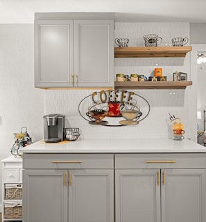 A kitchen with grey cabinets, a coffee station, and a white tiled backsplash.